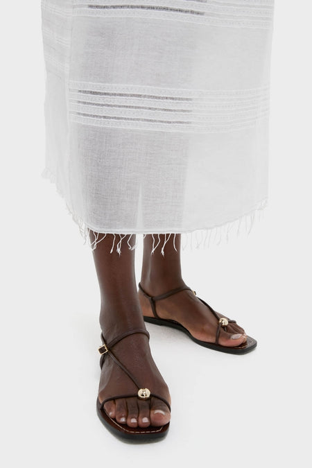 Close-up of lower legs in classic dark-brown T-strap sandals, showcasing the Abira White Nia Slip Dress's effortless, high-end woven pattern with hand-twisted fringe hem.