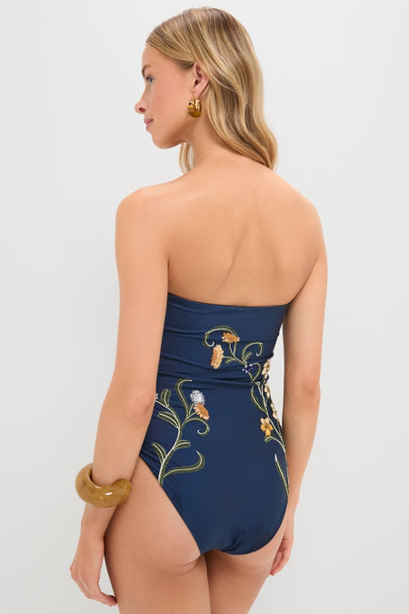 Woman wearing Laguna Durazno Embroidered One Piece, a classic, high-end strapless navy swimsuit with floral vines, accessorized with gold jewelry, epitomizing effortless elegance and fun.