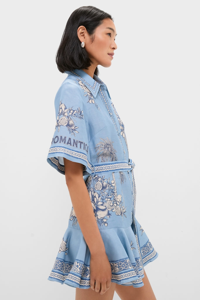 Model wearing the Blue Villa Romantica Mini Dress, a classic powder-blue romper with a fun floral and fruit print, belted waist, and high-end ruffled hem.