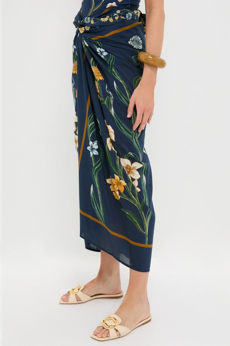 Laguna Lavanda Midi Sarong in navy-blue with floral print, knotted at waist, paired with cream slide sandals and amber bangle, exuding effortless, high-end style.