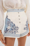 Woman wearing Cream Villa Romantica Corset Shorts with bird and foliage print, high-rise fit, raffia buttons, and pockets; an effortless, high-end style for warm days.