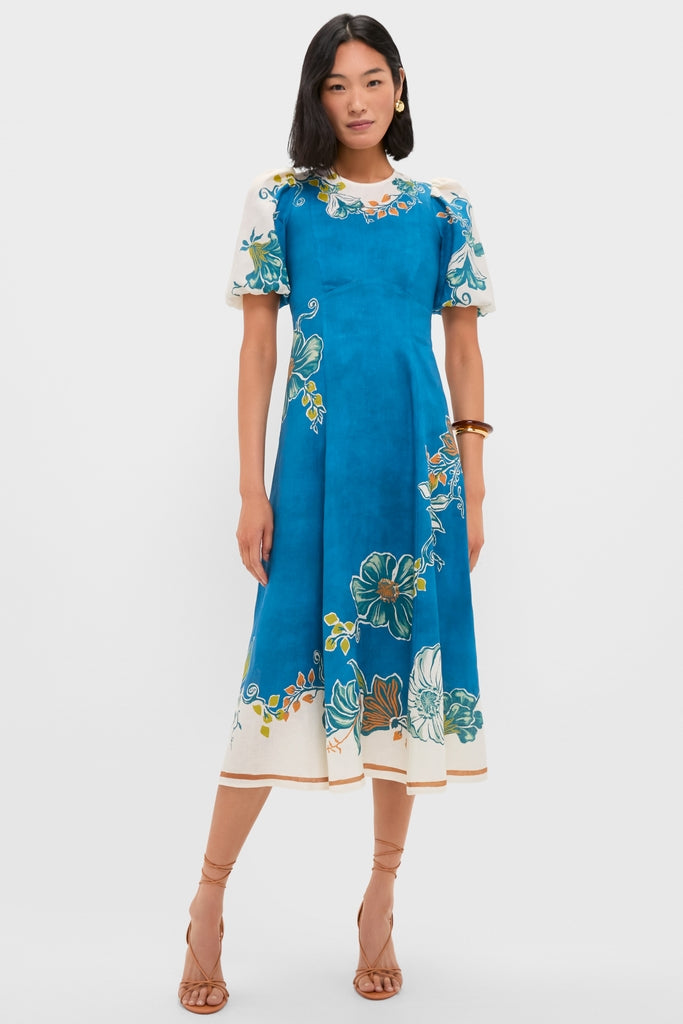 Woman wearing Blue Luda Midi Dress with painterly floral motifs, puff sleeves, and tan sandals. A classic, effortless, high-end look perfect for fun, warm-weather occasions.