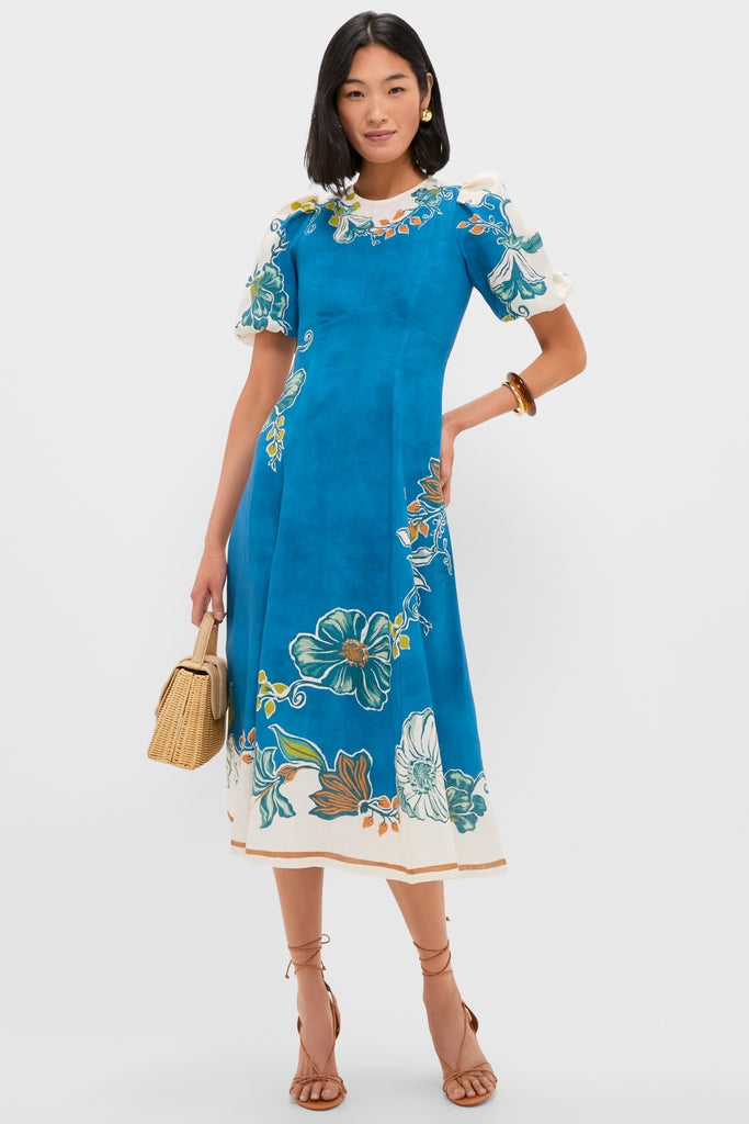 Woman models the Blue Luda Midi Dress, an effortless, classic piece with puff sleeves, floral print, and wicker handbag, embodying fun, high-end style.