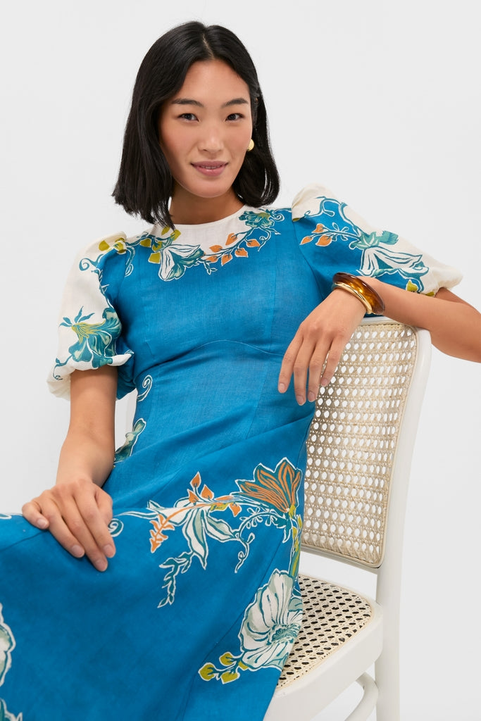 Young woman in Blue Luda Midi Dress with floral motifs, gold hoop earring, and amber bangle, embodies effortless, high-end style for classic, fun warm weather outings.