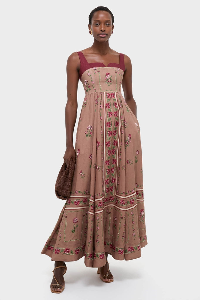 Woman in Quebrada Jaspe Maxi Dress featuring classic empire waist, floral embroidery, and square neckline. Effortless style with woven clutch and strappy sandals, exuding high-end, fun elegance.