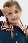 Young girl smiling in a Mariner Collar Cardigan in Navy, featuring a playful salute. This classic, high-end piece embodies effortless, fun style with embroidered boat motifs.