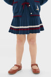 Child wearing the Pleated Knit Skirt in Navy, featuring classic pleats, burgundy stripe trim, and a fun bow detail. Effortless, high-end style with matching cardigan visible.
