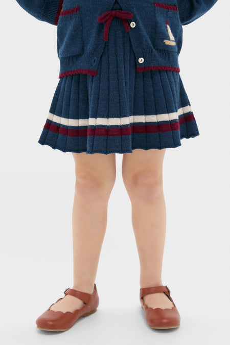 Child wearing the Pleated Knit Skirt in Navy, featuring classic pleats, burgundy stripe trim, and a fun bow detail. Effortless, high-end style with matching cardigan visible.