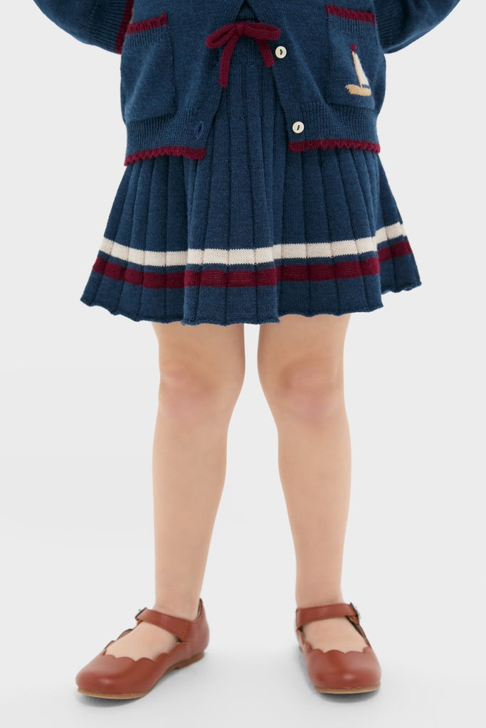 Child wearing the Pleated Knit Skirt in Navy, featuring classic pleats, burgundy stripe trim, and a fun bow detail. Effortless, high-end style with matching cardigan visible.