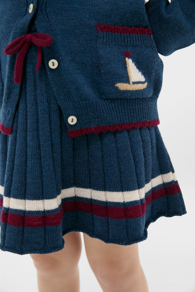 Child wearing Pleated Knit Skirt in Navy and matching cardigan, showcasing classic and effortless style with fun burgundy bow and high-end craftsmanship.