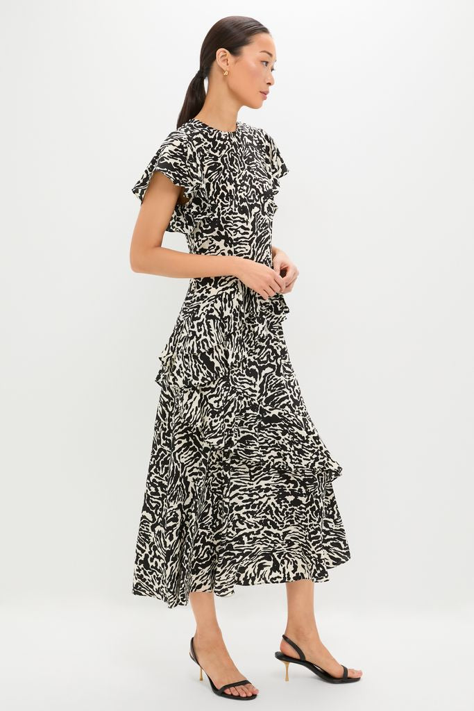 Woman modeling the Spotted Jasper Amaia Dress, featuring an effortless black-and-white abstract print, flutter sleeves, and tiered ruffles, paired with classic black slingback sandals.