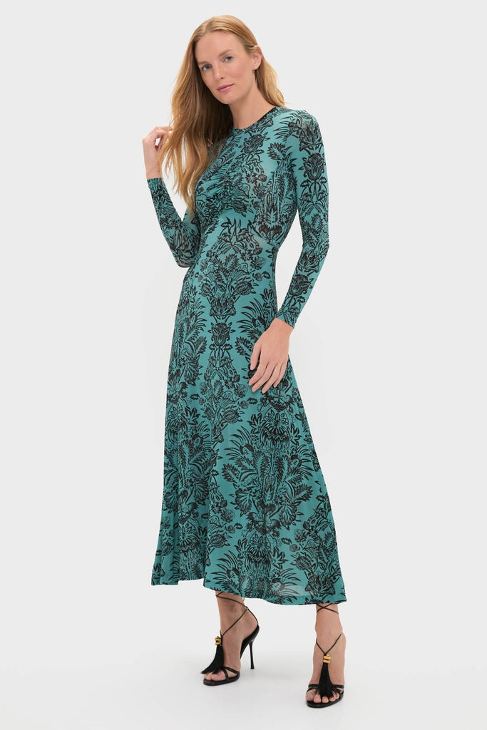 Model wearing the Ocean Flora Naia Dress, a classic, ankle-length teal dress with baroque floral patterns, high neckline, and flared skirt. Effortless style with strappy sandals.