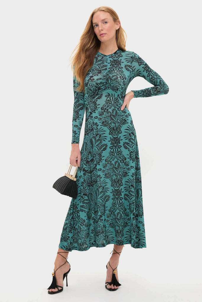 Woman in Ocean Flora Naia Dress, a classic, ankle-length teal piece with a black floral pattern, holding a black woven handbag, and wearing strappy, high-end sandals.