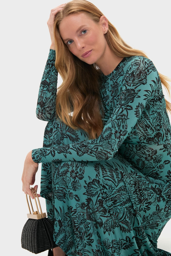 Woman in a classic Ocean Flora Naia Dress, featuring an effortless teal floral pattern, holds a fun, high-end black woven bag with gold-tone handles.