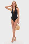 Young woman in Noir Milos Maillot, a classic black halter swimsuit with stone-embellished ring, holding a straw hat, exuding effortless, high-end style.