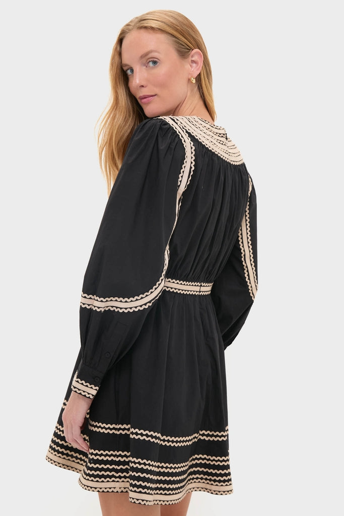 Young woman in a classic Noir Fiora Mini Dress with cream wavy trim, showcasing effortless style and fun elegance against a light backdrop.