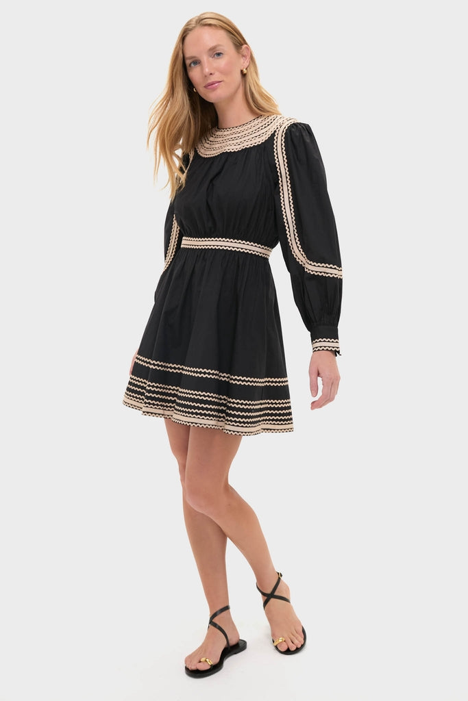Young woman in an effortlessly classic Noir Fiora Mini Dress with scalloped trim, paired with high-end black sandals, embodying fun and confidence against a neutral backdrop.