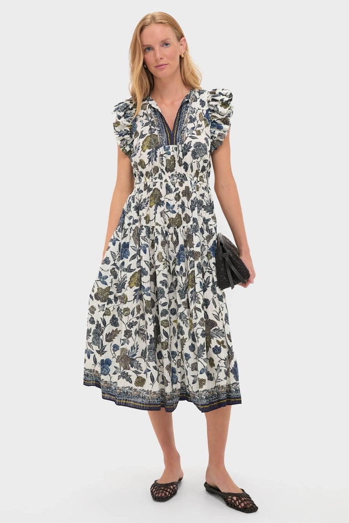 Woman models the Porcelain Kairi Dress, a classic, high-end, mid-length floral dress with ruffled cap sleeves and a tiered skirt, paired with a black clutch and flats.
