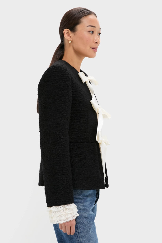 Woman in profile wearing the Black Seraphine Solid Bow Jacket with ivory ribbon bows and lace cuffs, paired with blue jeans, exuding an effortless, classic style.