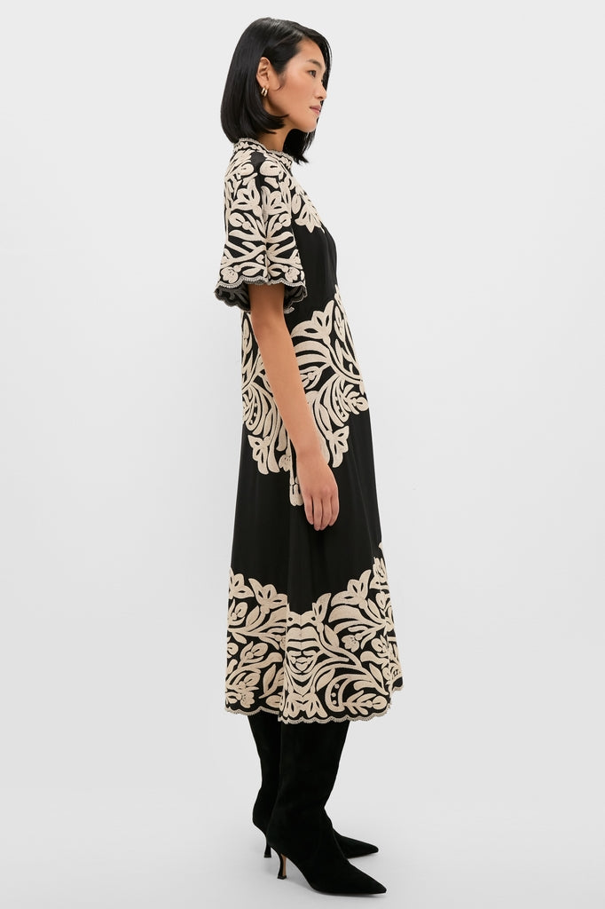 Side profile of a woman in the Black Alexandra Embroidery Short Sleeve Midi Dress with scrolling beige floral details, paired effortlessly with knee-high suede boots for a classic, high-end look.