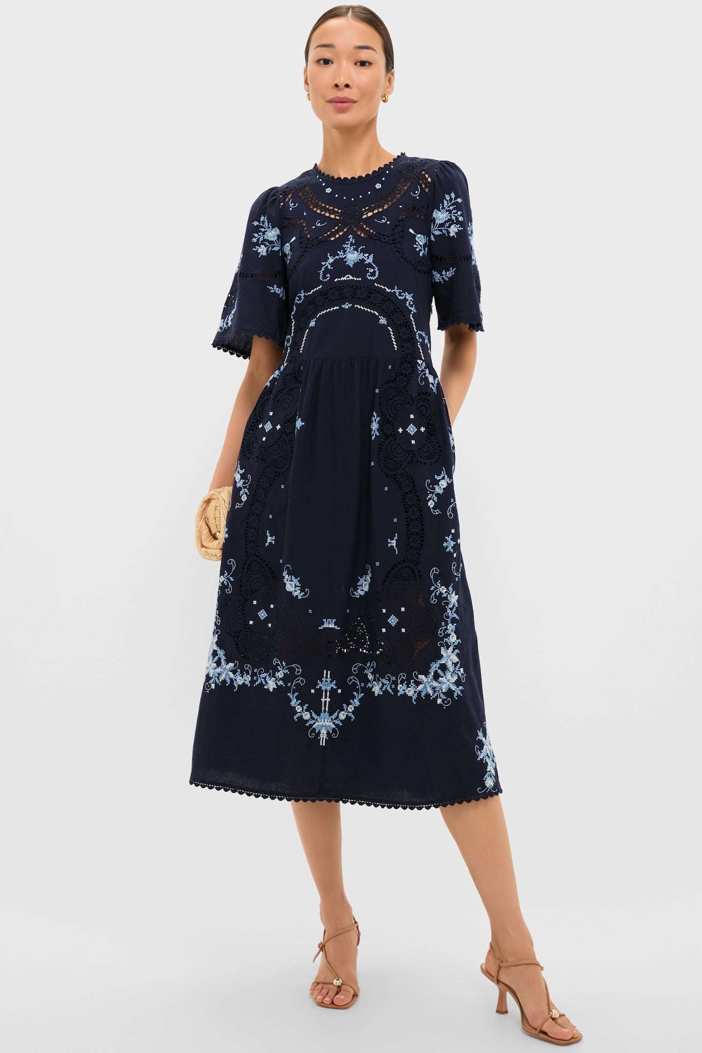 Exclusive Navy Edwina Embroidery Midi Dress features classic floral embroidery, intricate lace insets, and effortless bell sleeves, styled with a fun straw clutch and high-end sandals.
