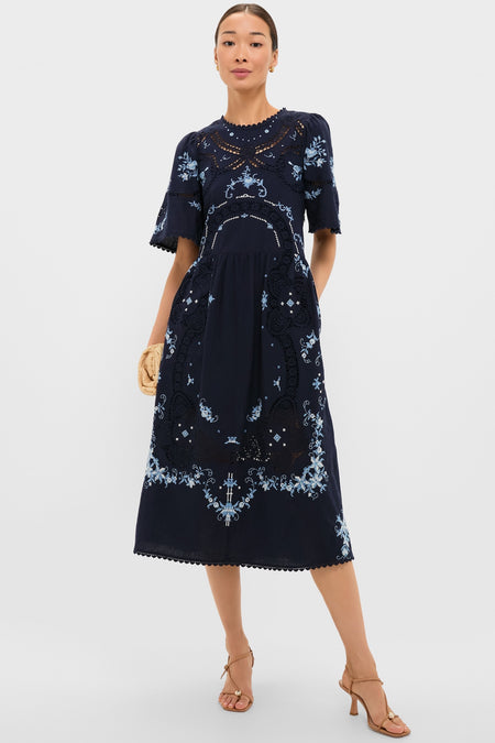 Exclusive Navy Edwina Embroidery Midi Dress features classic floral embroidery, intricate lace insets, and effortless bell sleeves, styled with a fun straw clutch and high-end sandals.