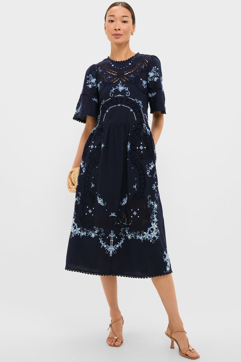Exclusive Navy Edwina Embroidery Midi Dress features classic floral embroidery, intricate lace insets, and effortless bell sleeves, styled with a fun straw clutch and high-end sandals.