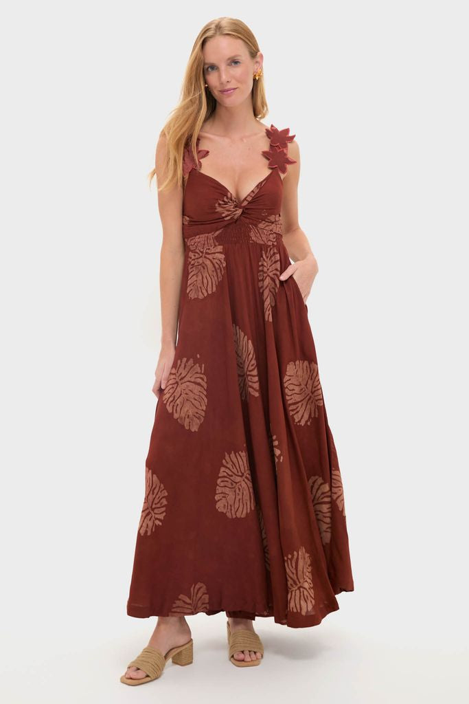 Woman models the Rust Toye Dress, a classic, high-end maxi with a knotted bust, appliqué leaf straps, smocked empire waist, and tropical print, embodying effortless and fun style.