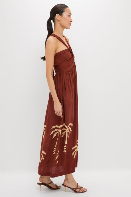 Model in a classic Rust Anike Dress featuring a twisted knot detail and palm-tree print, paired with gold earrings and strappy sandals, exuding an effortless, high-end style.