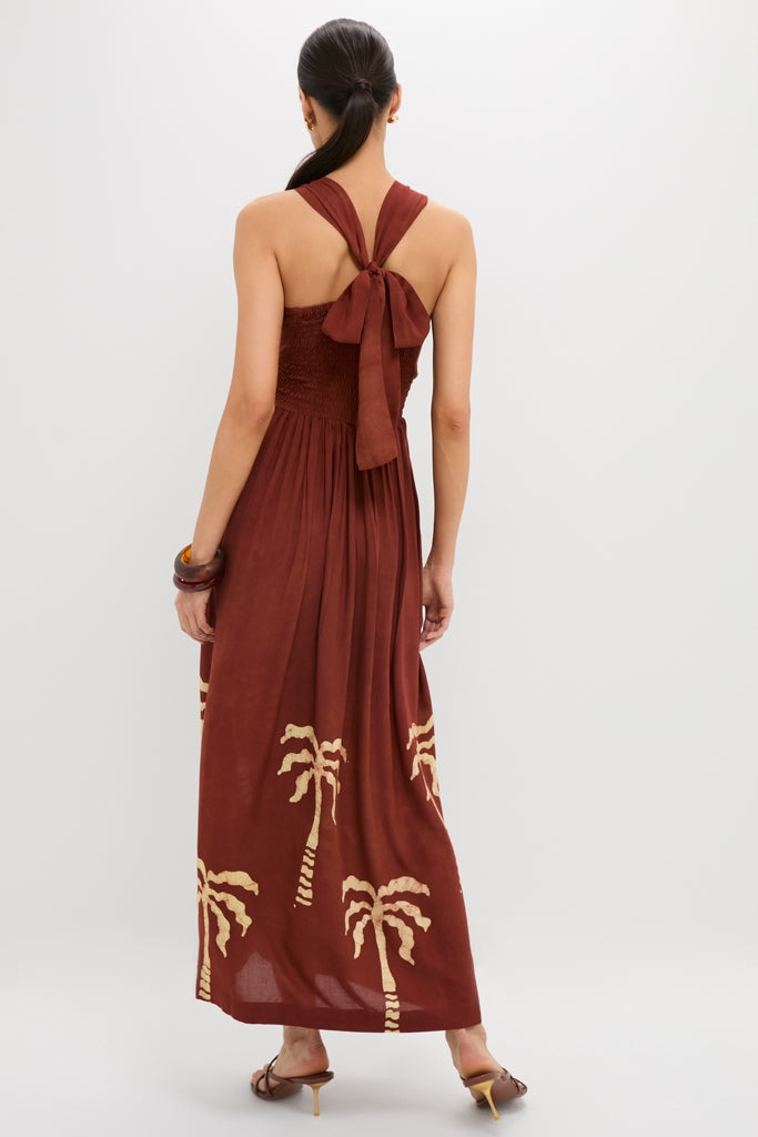 Woman in a Rust Anike Dress with palm tree print, showcasing an effortless, classic style. The halter-neck ties into a bow, paired with high-end heeled sandals.