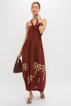 Model wears the Rust Anike Dress, a classic halter-neck maxi with palm-tree motif, accessorized with a woven handbag, chunky bangles, and sandals for an effortless, high-end look.