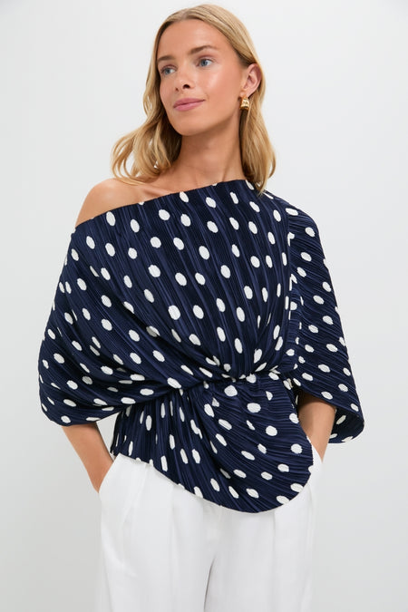 Woman models the Small Dot Deep Lake Cami Top, a classic, one-shoulder navy pleated blouse with large white polka dots, paired effortlessly with high-waisted trousers for a fun, high-end look.