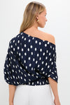 Woman in a classic Small Dot Deep Lake Cami Top, an effortless navy-blue, one-shoulder blouse with white polka dots, paired with high-waisted pants, exuding high-end fun.