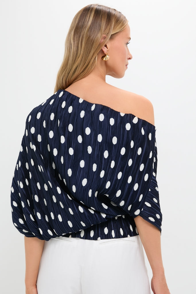 Woman in a classic Small Dot Deep Lake Cami Top, an effortless navy-blue, one-shoulder blouse with white polka dots, paired with high-waisted pants, exuding high-end fun.