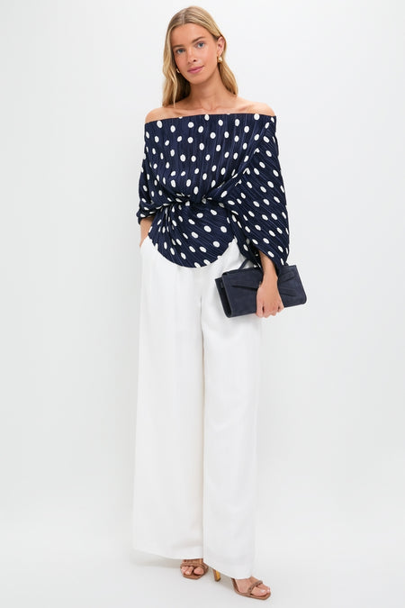 Model wearing the Small Dot Deep Lake Cami Top, an off-the-shoulder navy blouse with white polka dots, paired with classic white trousers and a structured navy clutch for an effortless, high-end look.