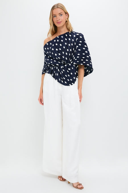 Model wearing the Small Dot Deep Lake Cami Top, a classic navy polka dot one-shoulder blouse, paired with effortless high-waisted white trousers and tan sandals.