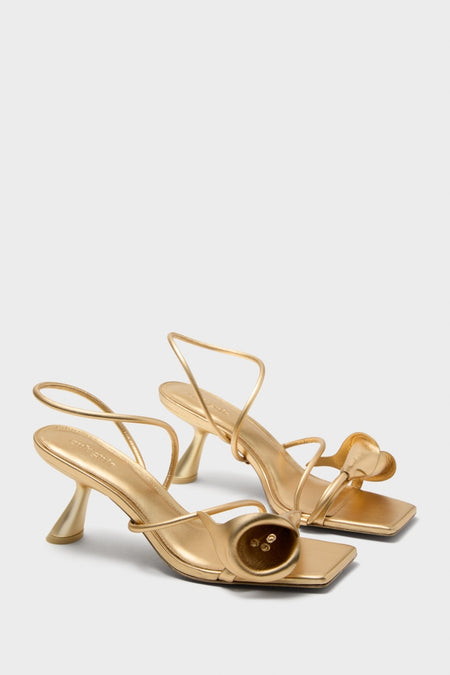 Brass Ria Sandals: A classic, high-end pair with metallic gold finish, featuring square toe, looping straps, sculptural flared heels, and a decorative button detail. Effortlessly fun and stylish.