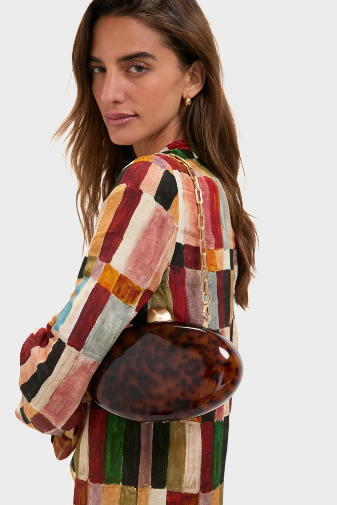 Woman in a multicolored dress holds the Caramel Tortoise Eviana Clutch, a classic, high-end acrylic purse with a gold-tone clasp and chain, exuding effortless sophistication.