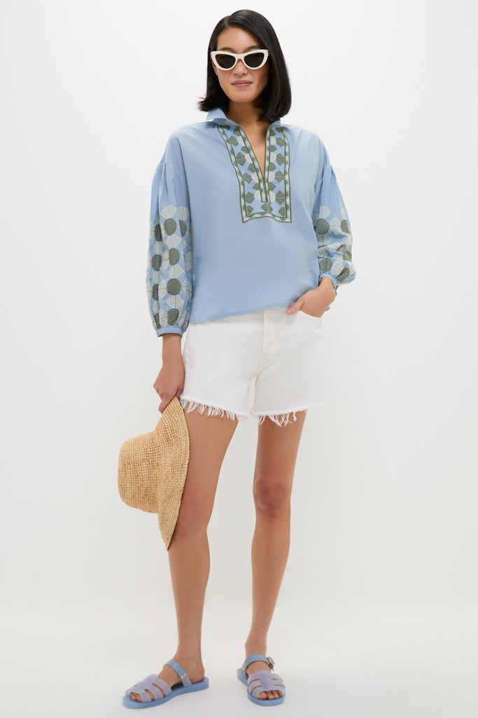 Woman in Sky Geometric Embroidered Paget Top, embodying classic, effortless style with blouson sleeves and embroidery, paired with shorts and sandals, holding a straw hat.