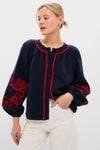 Woman models the Navy & Red Soutache Stella Cardigan, a classic navy button-front with red trim, playful squiggle detailing, paired effortlessly with high-waisted pants.