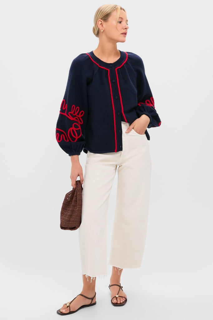 Woman models the Navy & Red Soutache Stella Cardigan, featuring bold red piping and embroidery. Paired with cream wide-leg cropped jeans and black sandals for an effortless, high-end look.