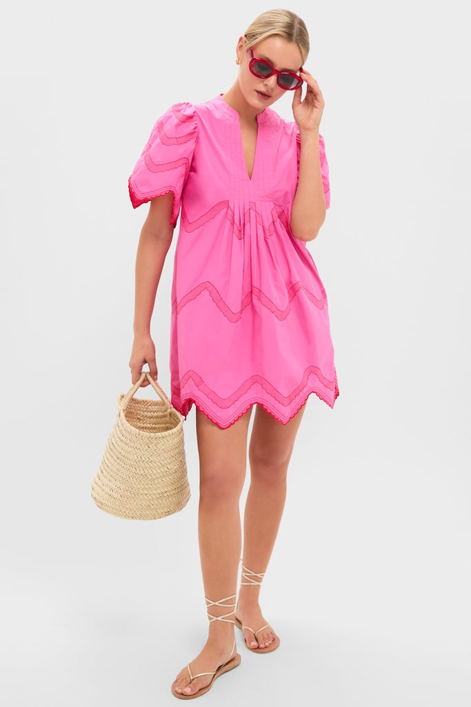 Woman in a hot pink Weston Mini Dress with red embroidery, scalloped hem, and wave detailing, styled with classic strappy sandals and a woven basket bag for an effortless, high-end look.