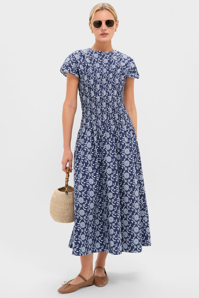 Model wears the classic Blue Floral Smocked Rollins Midi Dress with effortless cap sleeves, paired with fun tortoiseshell sunglasses, tan ballet flats, and a high-end straw bucket bag.