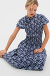 Young woman in a classic, high-end Blue Floral Smocked Rollins Midi Dress, featuring an effortless smocked bodice and fun white floral print, seated gracefully.