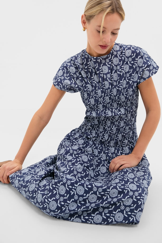 Young woman in a classic, high-end Blue Floral Smocked Rollins Midi Dress, featuring an effortless smocked bodice and fun white floral print, seated gracefully.
