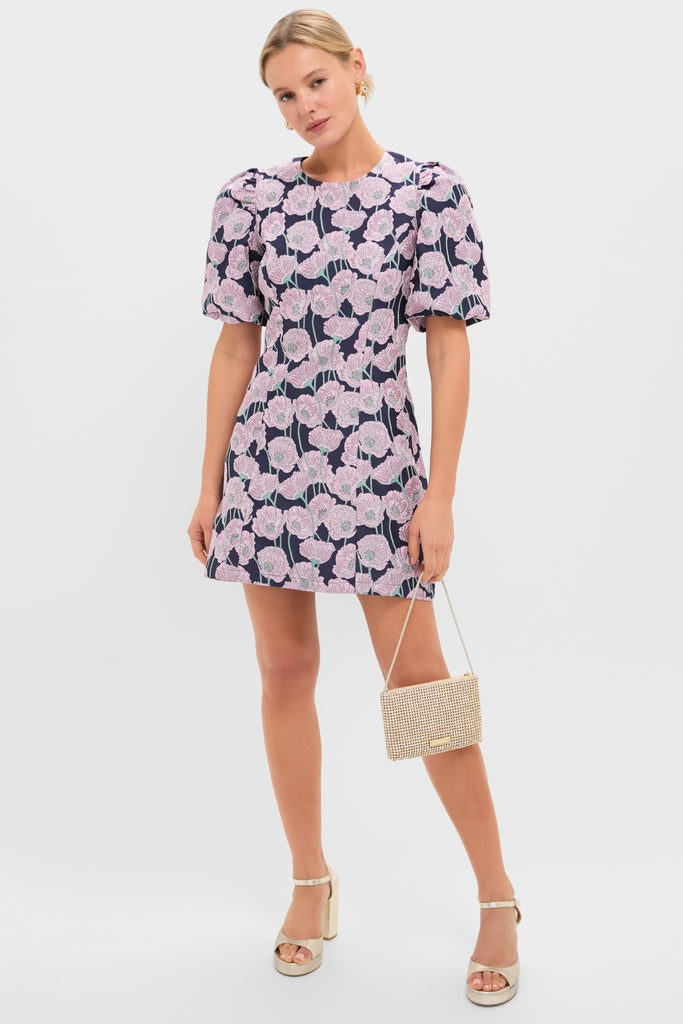 Model in Pink & Navy Floral Jacquard Minetta Mini Dress, showcasing its classic, effortless style with puffed sleeves and a high-end finish, paired with a fun clutch and platform sandals.