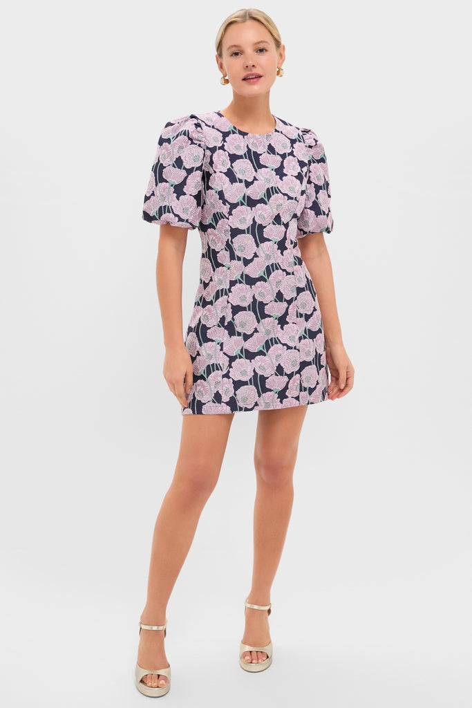 Young woman models the Pink & Navy Floral Jacquard Minetta Mini Dress, featuring classic puff sleeves, effortlessly styled with ankle-strap heels and gold earrings for a fun, high-end look.