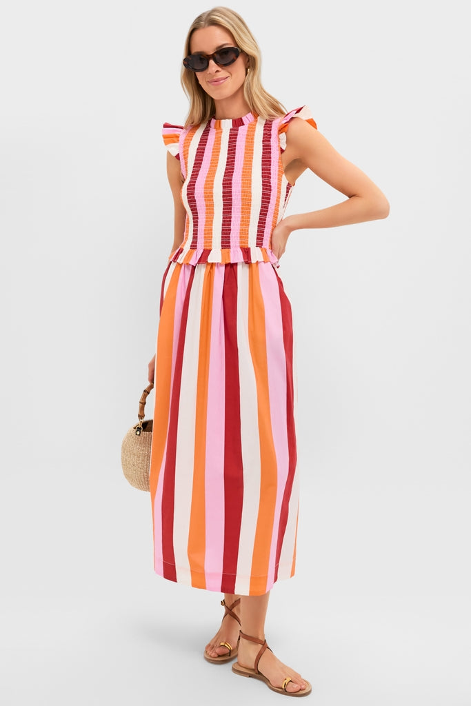 Woman models the Orange, Pink & Crimson Stripe Smocked Sully Dress with classic smocked bodice, flutter sleeves, accessorized with a straw handbag, embodying high-end, effortless fun.