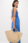 Woman in classic cobalt-blue dress holds the Natural Mimi Basket, a high-end, effortless raffia tote with ring handles and gold charms, exuding fun elegance.