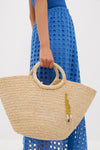 Person holding the Natural Mimi Basket, a classic raffia tote with round handles and a gold-beaded charm, paired with a blue eyelet dress for an effortless, high-end look.